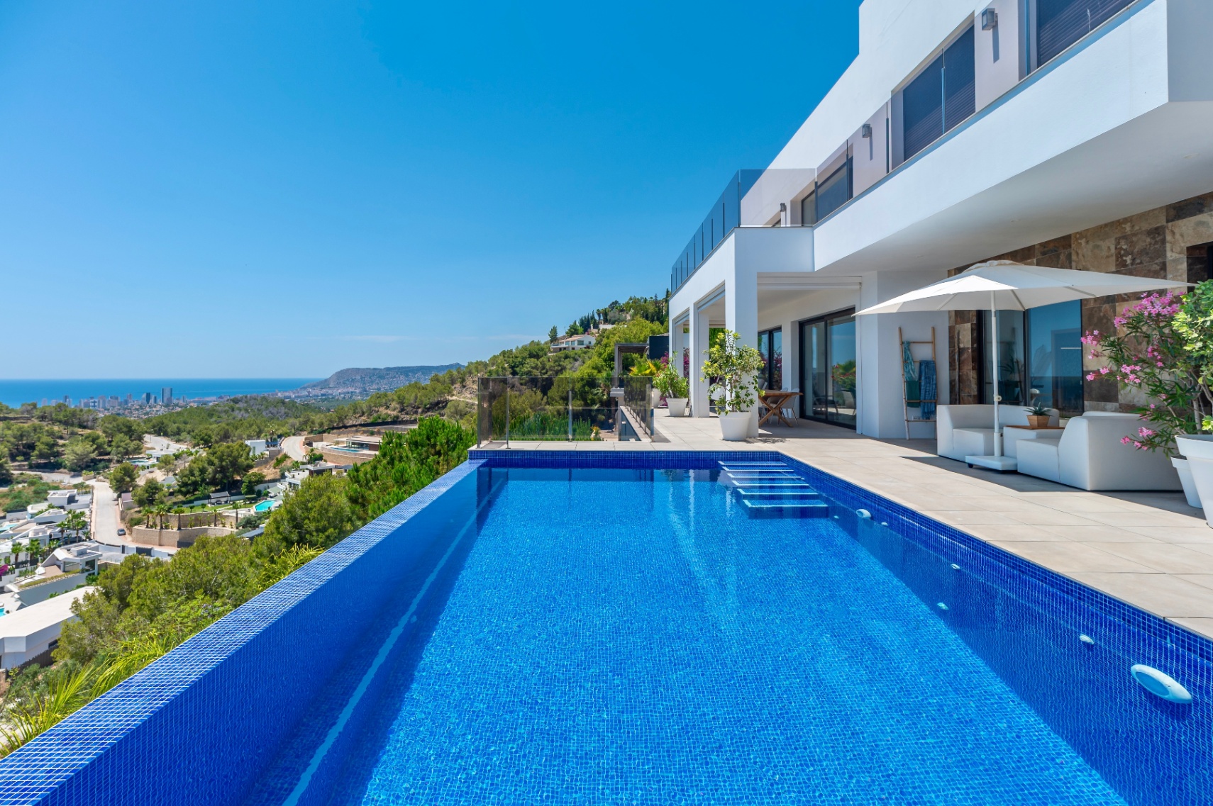 Piscine à débordement en mosaïque bleue avec marches intégrées, jouxtant une terrasse spacieuse avec mobilier de salon et vues panoramiques sur la côte et la ville. Piscine à débordement en mosaïque bleue avec marches intégrées, jouxtant une terrasse spacieuse avec mobilier de salon et vues panoramiques sur la côte et la ville.