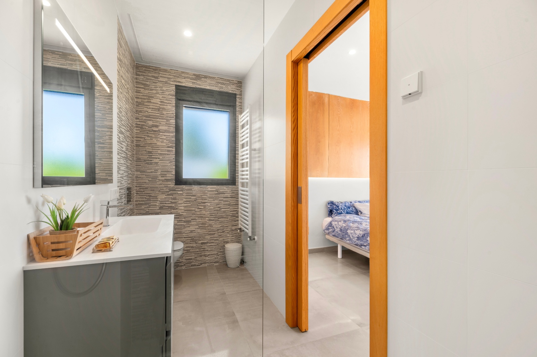 Salle de bain contemporaine avec meuble-lavabo gris, miroir éclairé et carrelage mural texturé. Une fenêtre dépolie et un sèche-serviettes blanc sont présents, avec accès à une chambre. Salle de bain contemporaine avec meuble-lavabo gris, miroir éclairé et carrelage mural texturé. Une fenêtre dépolie et un sèche-serviettes blanc sont présents, avec accès à une chambre.