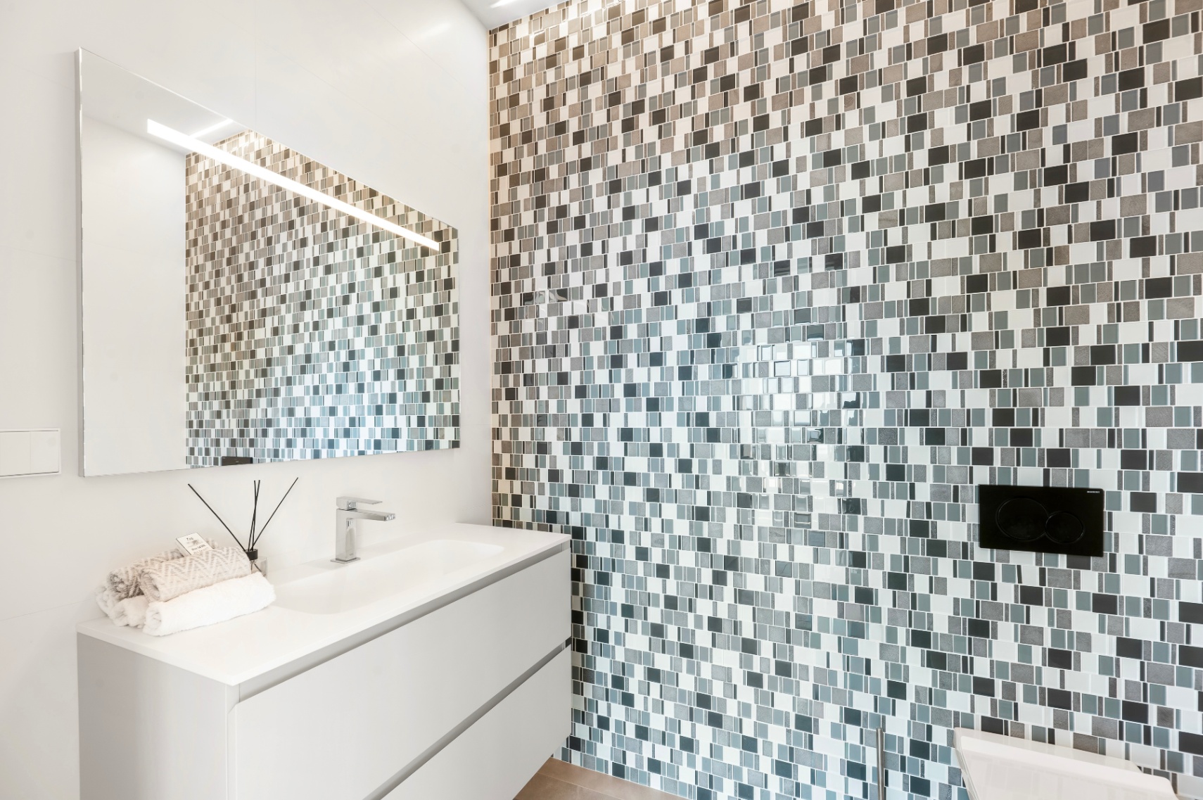 Salle de bain moderne avec meuble vasque suspendu blanc et lavabo intégré. Le mur est revêtu d'une mosaïque de carreaux gris et blancs, rehaussée par un grand miroir lumineux. Salle de bain moderne avec meuble vasque suspendu blanc et lavabo intégré. Le mur est revêtu d'une mosaïque de carreaux gris et blancs, rehaussée par un grand miroir lumineux.