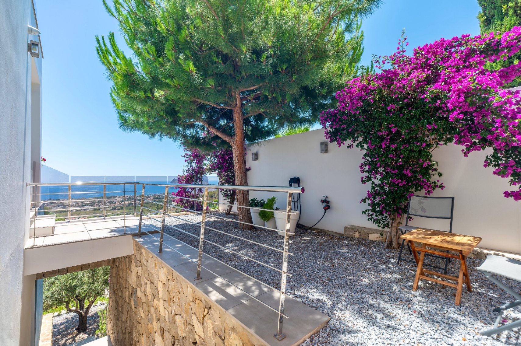 Terrasse avec garde-corps en acier inoxydable et verre, offrant une vue panoramique sur la mer. Jardin de gravier adjacent avec un grand pin et une bougainvillée violette sur un mur blanc. Terrasse avec garde-corps en acier inoxydable et verre, offrant une vue panoramique sur la mer. Jardin de gravier adjacent avec un grand pin et une bougainvillée violette sur un mur blanc.