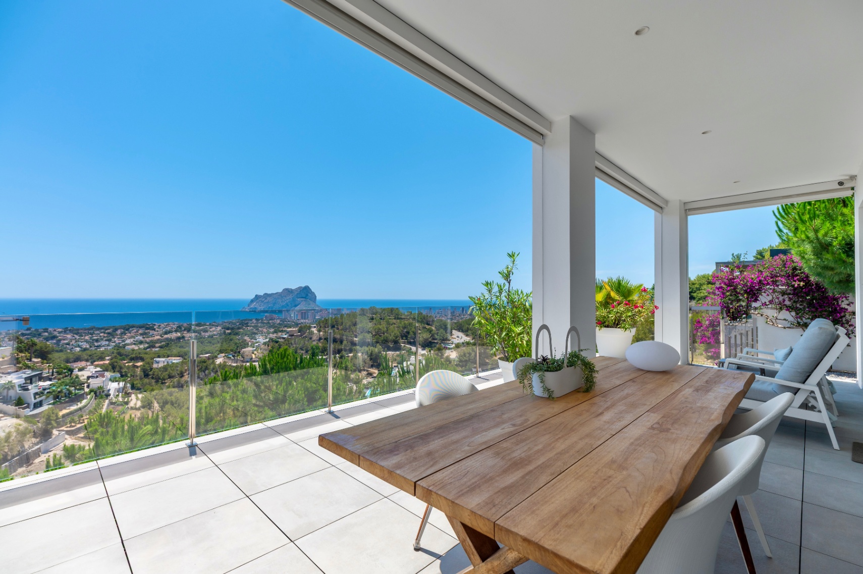 Terrasse couverte spacieuse avec une table à manger en bois massif et des chaises modernes blanches. Vues panoramiques sur la mer, Calpe et le Peñón de Ifach derrière une balustrade en verre. Terrasse couverte spacieuse avec une table à manger en bois massif et des chaises modernes blanches. Vues panoramiques sur la mer, Calpe et le Peñón de Ifach derrière une balustrade en verre.