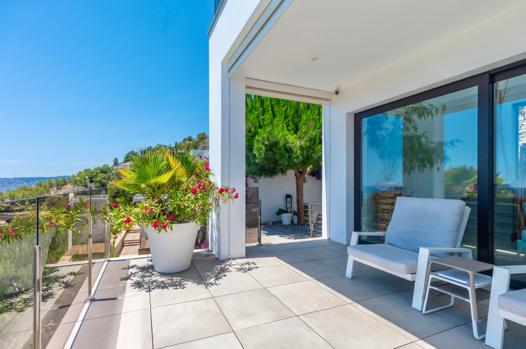 Terrasse extérieure moderne avec carrelage gris clair et garde-corps en verre, offrant une vue dégagée sur la colline verdoyante. Aménagée avec des sièges et des plantes en pot sous un soleil éclatant. Terrasse extérieure moderne avec carrelage gris clair et garde-corps en verre, offrant une vue dégagée sur la colline verdoyante. Aménagée avec des sièges et des plantes en pot sous un soleil éclatant.
