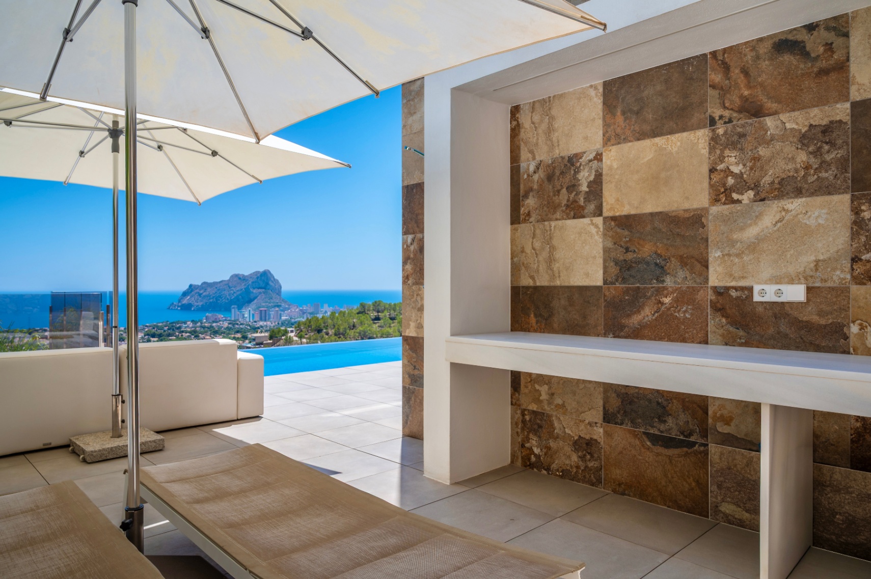 Vaste terrasse extérieure avec comptoir intégré et carrelage mural effet pierre naturelle aux tons variés. Vue panoramique sur la mer, la piscine à débordement et le Peñon de Ifach. Vaste terrasse extérieure avec comptoir intégré et carrelage mural effet pierre naturelle aux tons variés. Vue panoramique sur la mer, la piscine à débordement et le Peñon de Ifach.