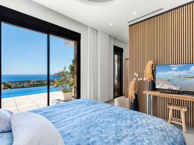 Chambre avec de grandes portes coulissantes en verre s'ouvrant sur une piscine à débordement et des vues panoramiques sur la mer. Un mur moderne en lattes de bois encadre la télévision, complétant l'intérieur clair.