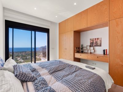 Chambre principale avec de grandes baies vitrées coulissantes offrant une vue panoramique sur la mer et la côte. Armoires intégrées en bois clair avec bureau.