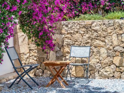 Coin salon extérieur avec deux chaises pliantes grises et une table en bois sur gravier. Un mur en pierre naturelle est orné de bougainvilliers magenta éclatants.