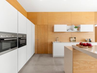 Cuisine moderne avec armoires blanches intégrées et fours Bosch. L'îlot central en pierre claire et les panneaux muraux et plafond en bois clair caractérisent l'espace.