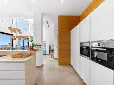 Cuisine moderne épurée avec fours Bosch intégrés, armoires blanches sans poignées et îlot en pierre claire. L'agencement ouvert s'étend vers la salle à manger avec vue panoramique sur la mer.