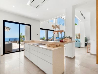 Cuisine ouverte moderne avec îlot central en comptoir beige et plaque à induction intégrée. De larges baies vitrées coulissantes offrent une vue directe sur la mer Méditerranée, éclairant l'espace contemporain.