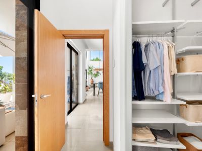 Entrée de couloir moderne avec porte en bois clair ouverte, révélant un placard blanc intégré avec étagères modulables et un aperçu de terrasse ensoleillée avec vue sur mer.