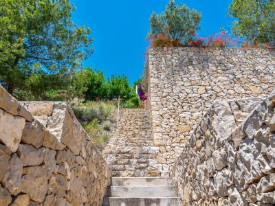 Escalier extérieur en pierre naturelle bordé de murs en pierres sèches, s'élevant au milieu d'une végétation méditerranéenne luxuriante sous un ciel azur.