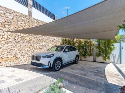 Espace de stationnement extérieur abrité par une voile d'ombrage, avec une allée pavée bicolore. Un mur en pierres sèches et des jardinières végétalisées encadrent la zone.