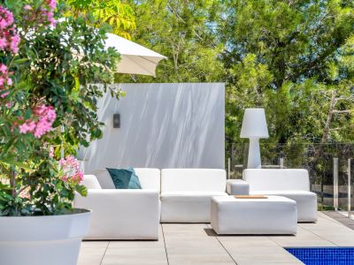 Espace lounge extérieur contemporain avec canapés modulaires blancs et pouf assorti sur une terrasse carrelée. Bordé par une rambarde en verre, une végétation luxuriante et une plante à fleurs roses.