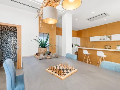 Espace salle à manger contemporain avec table grise et chaises bleues. Un lustre en bois naturel et une cuisine ouverte avec boiseries claires et comptoir sont visibles. Accès à une salle de bain carrelée.