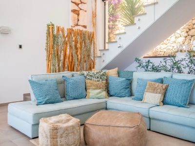 Espace salon chaleureux avec canapé sectionnel bleu clair, coussins variés et deux poufs en cuir. Escalier moderne en verre près d'un écran en bois naturel et plantes, mur de pierre visible.
