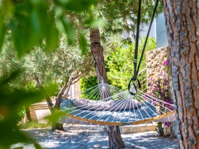 Hamac gris rembourré avec coussin, suspendu entre des arbres feuillus sur un sol de gravier. Un mur en pierre avec des fleurs violettes orne l'arrière-plan du jardin.