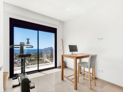 Pièce polyvalente avec bureau haut en bois et vélo d'appartement, offrant des vues imprenables sur la mer et le Peñon de Ifach à travers les grandes baies vitrées coulissantes.