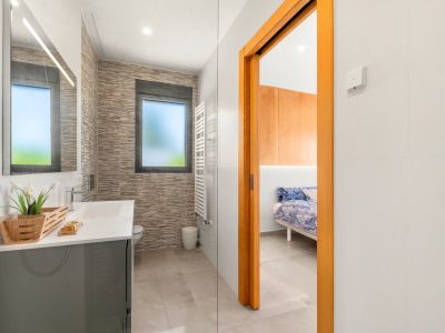 Salle de bain contemporaine avec meuble-lavabo gris, miroir éclairé et carrelage mural texturé. Une fenêtre dépolie et un sèche-serviettes blanc sont présents, avec accès à une chambre.