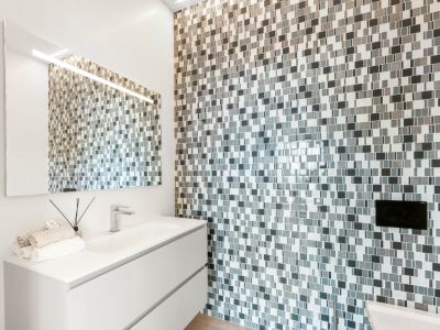 Salle de bain moderne avec meuble vasque suspendu blanc et lavabo intégré. Le mur est revêtu d'une mosaïque de carreaux gris et blancs, rehaussée par un grand miroir lumineux.
