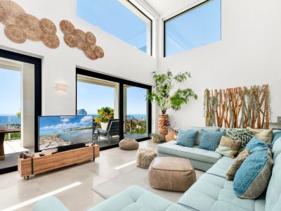 Salon lumineux avec de hauts plafonds et de grandes baies vitrées offrant une vue panoramique sur la mer. Un canapé d'angle bleu clair s'harmonise avec les éléments décoratifs en bois naturel.