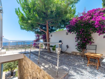 Terrasse avec garde-corps en acier inoxydable et verre, offrant une vue panoramique sur la mer. Jardin de gravier adjacent avec un grand pin et une bougainvillée violette sur un mur blanc.
