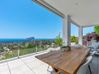 Terrasse couverte spacieuse avec une table à manger en bois massif et des chaises modernes blanches. Vues panoramiques sur la mer, Calpe et le Peñón de Ifach derrière une balustrade en verre.