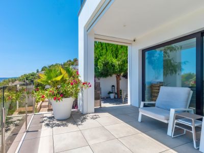 Terrasse extérieure moderne avec carrelage gris clair et garde-corps en verre, offrant une vue dégagée sur la colline verdoyante. Aménagée avec des sièges et des plantes en pot sous un soleil éclatant.