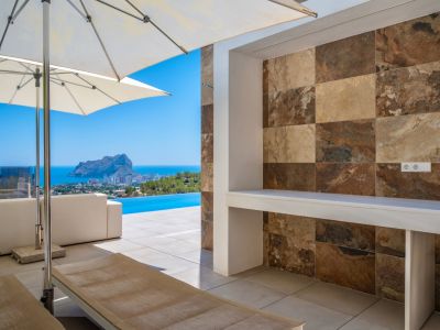 Vaste terrasse extérieure avec comptoir intégré et carrelage mural effet pierre naturelle aux tons variés. Vue panoramique sur la mer, la piscine à débordement et le Peñon de Ifach.