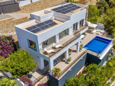 Vue aérienne d'une villa blanche moderne avec niveaux en terrasses, piscine rectangulaire et vastes espaces extérieurs. Panneaux solaires sur les toits plats, entourée d'un paysage méditerranéen luxuriant.