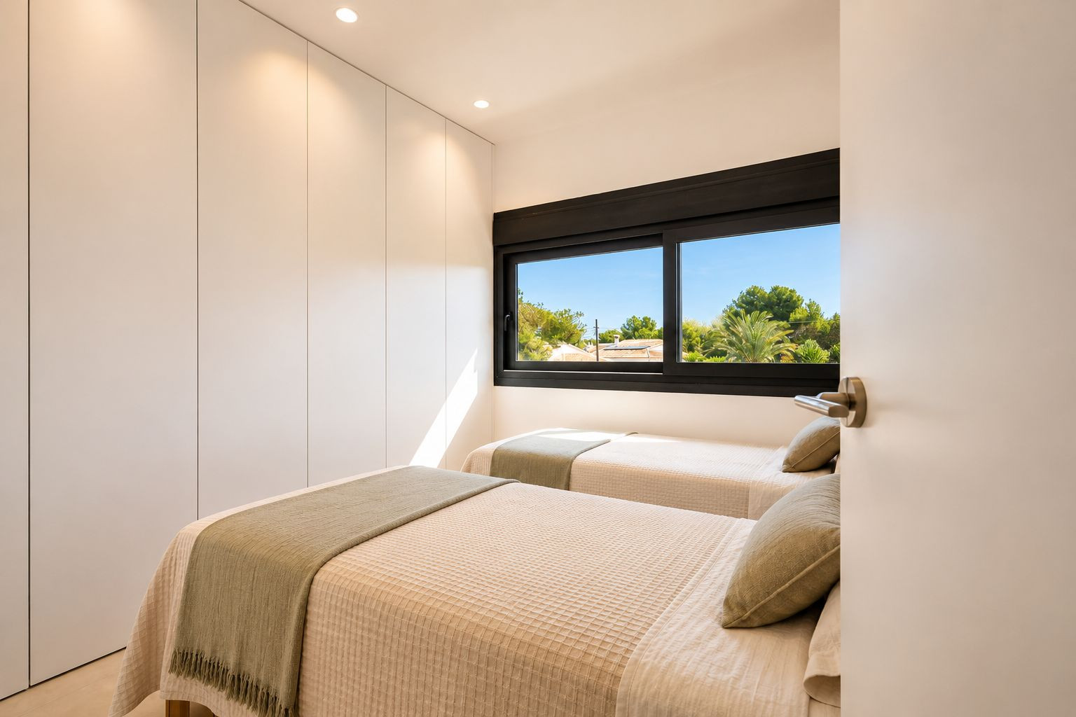 Chambre d'amis avec deux lits simples, placards intégrés blancs sans poignées et grande fenêtre offrant une vue sur la végétation méditerranéenne. Éclairage encastré et naturel. Chambre d'amis avec deux lits simples, placards intégrés blancs sans poignées et grande fenêtre offrant une vue sur la végétation méditerranéenne. Éclairage encastré et naturel.