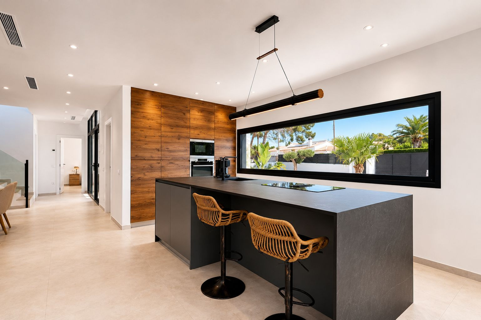 Cuisine moderne avec îlot central foncé intégrant plaque et évier, accompagnée de tabourets en rotin. Mur en bois avec électroménager encastré et large fenêtre sur le jardin. Cuisine moderne avec îlot central foncé intégrant plaque et évier, accompagnée de tabourets en rotin. Mur en bois avec électroménager encastré et large fenêtre sur le jardin.