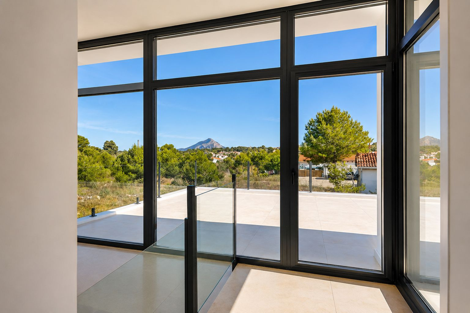 Grande baie vitrée à cadres sombres et balustrade en verre, reliant l'intérieur à une terrasse carrelée avec vue sur la pinède et la montagne. Grande baie vitrée à cadres sombres et balustrade en verre, reliant l'intérieur à une terrasse carrelée avec vue sur la pinède et la montagne.