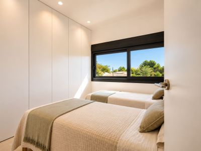 Chambre d'amis avec deux lits simples, placards intégrés blancs sans poignées et grande fenêtre offrant une vue sur la végétation méditerranéenne. Éclairage encastré et naturel.