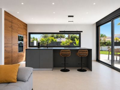 Cuisine moderne avec îlot central, équipée d'appareils intégrés et de tabourets de bar en rotin. De larges fenêtres offrent une vue sur le jardin et un accès direct à l'extérieur.