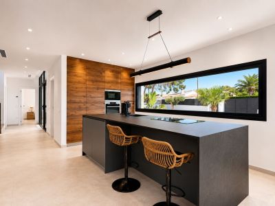 Cuisine moderne avec îlot central foncé intégrant plaque et évier, accompagnée de tabourets en rotin. Mur en bois avec électroménager encastré et large fenêtre sur le jardin.