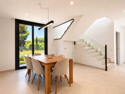 Espace repas moderne avec table en bois clair et six chaises, éclairé par une suspension linéaire noire. Une cage d'escalier contemporaine avec marches lumineuses et balustrade en verre y est attenante.