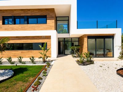 Façade de villa contemporaine avec revêtement effet bois, murs blancs et allée carrelée menant à l'entrée vitrée principale, complétée par un aménagement paysager méditerranéen minimaliste.