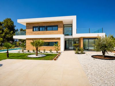 Vue extérieure de la villa moderne avec façade à deux étages, revêtement aspect bois et grandes baies vitrées. Allées carrelées, jardin paysager avec palmiers, olivier et piscine visible.