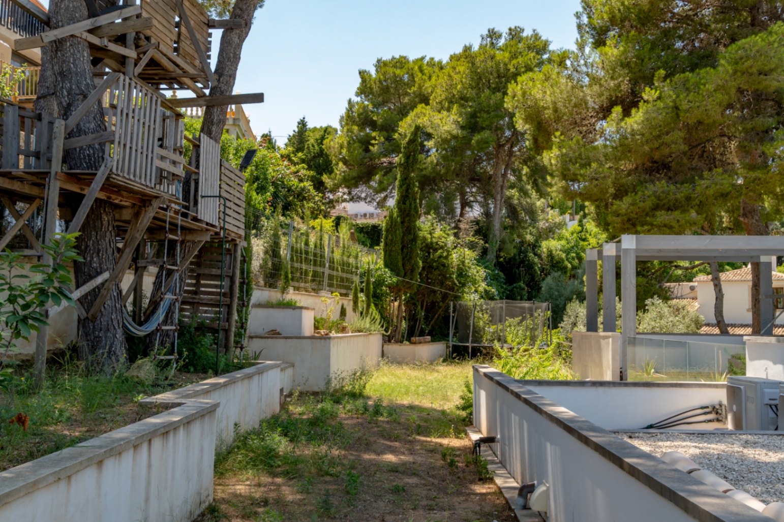 Cabane en bois rustique intégrée à des pins matures, avec un hamac. Le jardin en terrasses présente des murs en béton, une végétation dense et une pergola moderne.