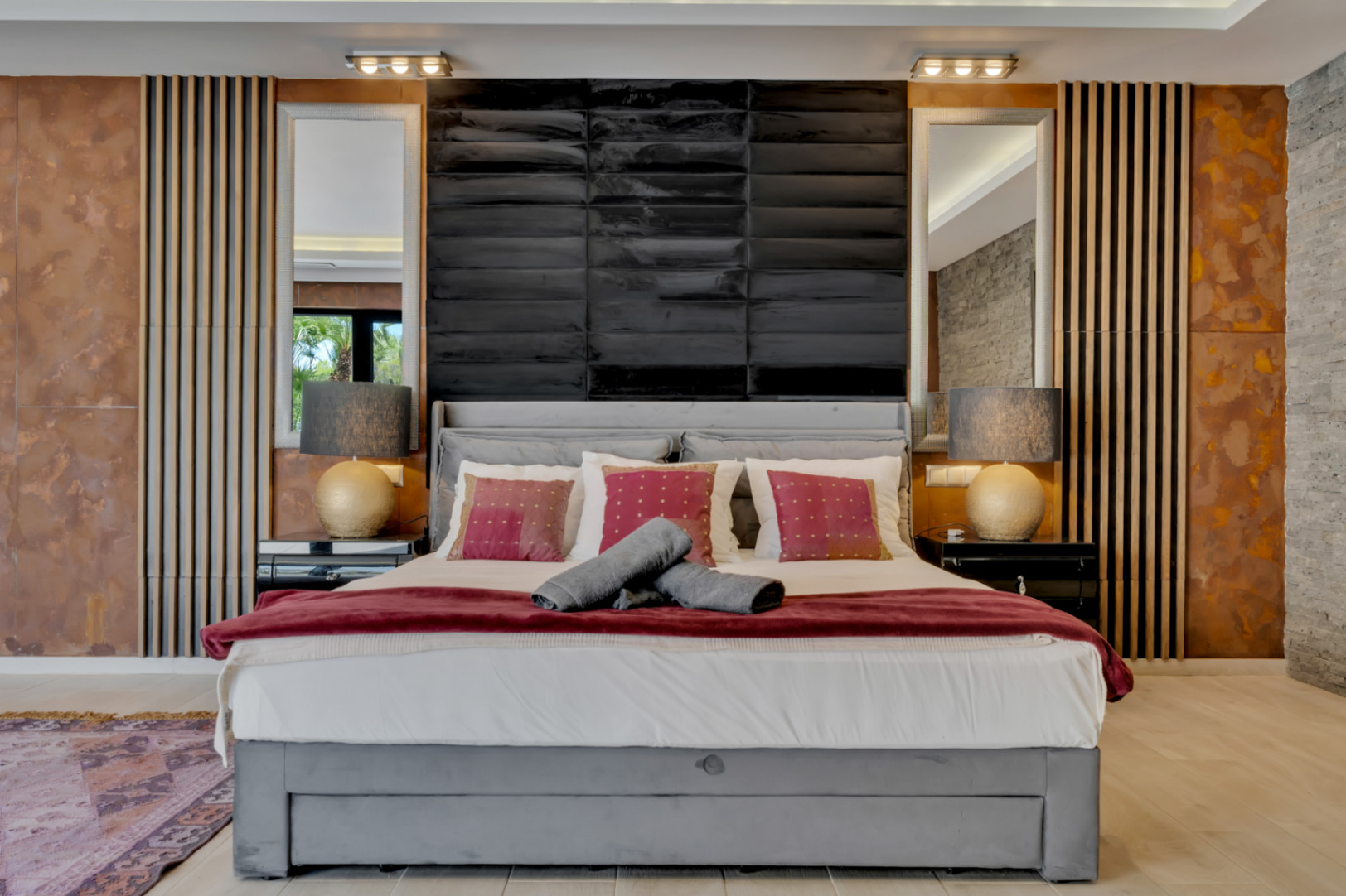 Chambre principale avec lit tapissé gris, tête de lit noire texturée, miroirs et panneaux muraux uniques en bois, métal vieilli et pierre. L'éclairage sur mesure crée une ambiance.