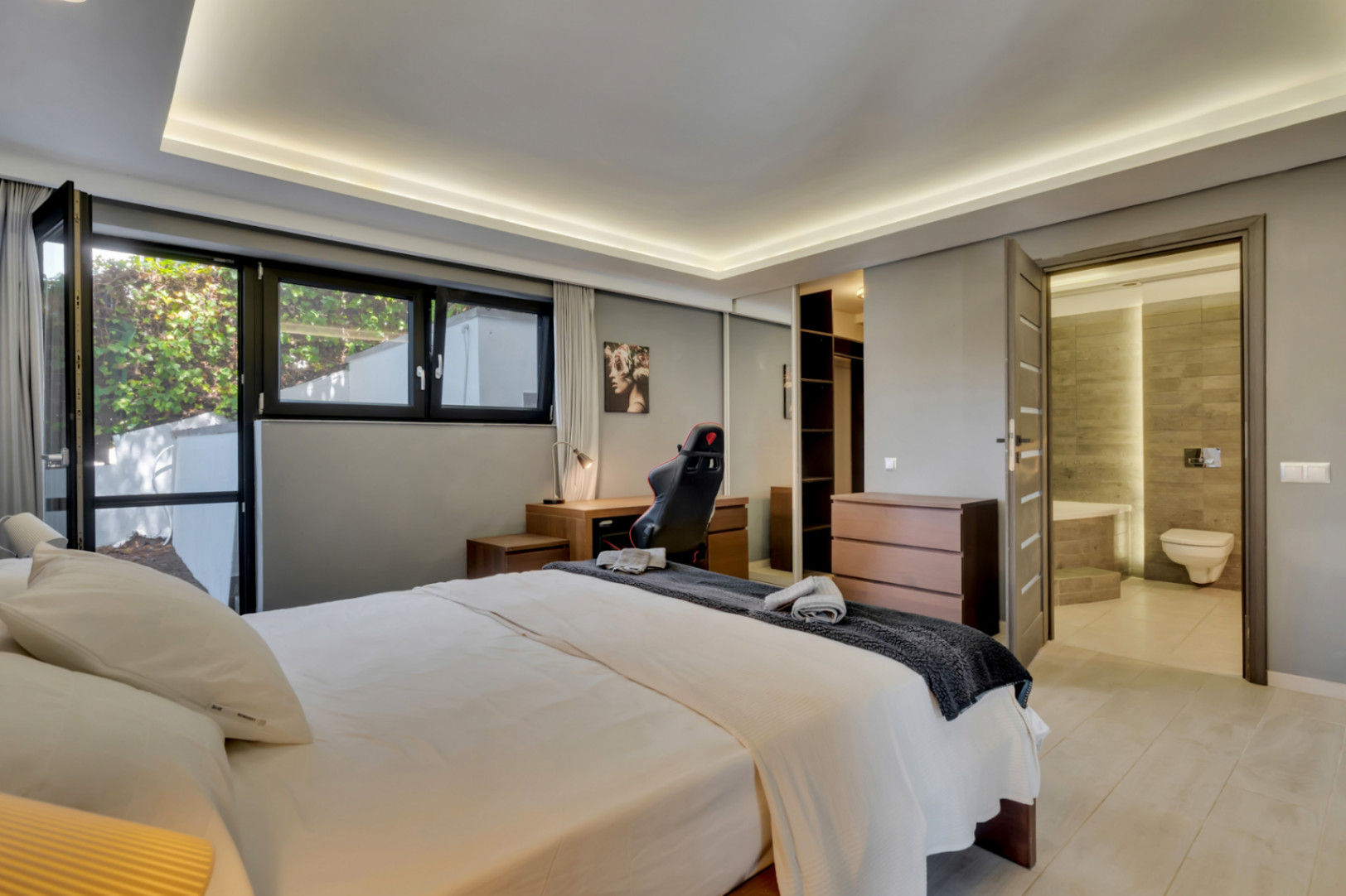 Chambre principale lumineuse avec lit double, bureau en bois et grandes fenêtres. Comprend une armoire à portes miroir, une commode et un accès direct à une salle de bain privative carrelée.