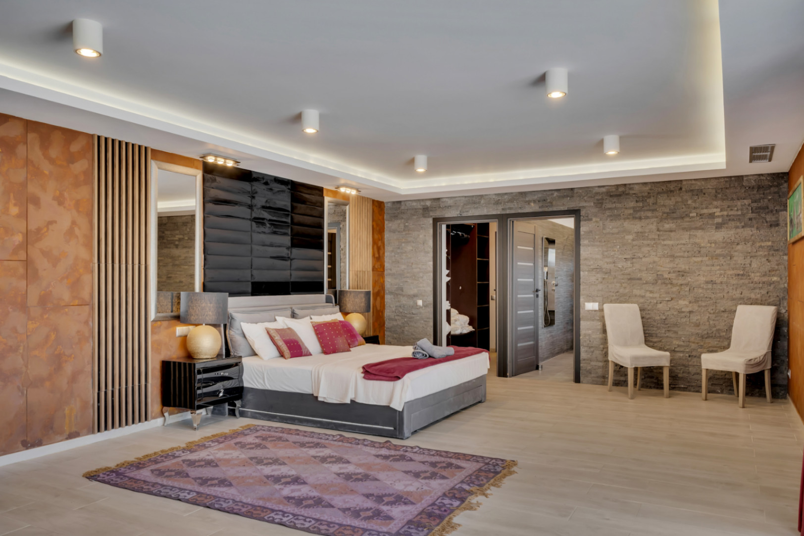 Chambre spacieuse au design moderne avec mur effet pierre texturé, tête de lit capitonnée et éclairage LED intégré. Accès direct à un dressing et une salle de bain attenante.