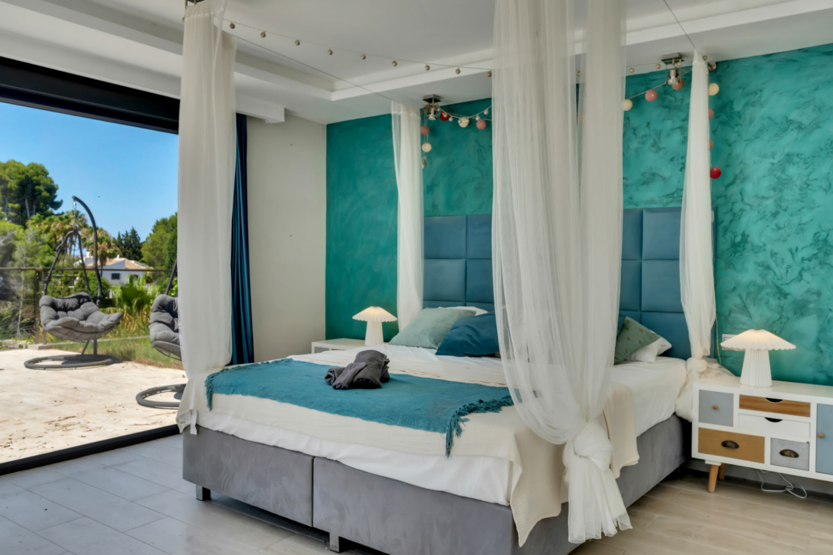 Chambre spacieuse avec lit à baldaquin, tête de lit bleue capitonnée, mur d'accent turquoise texturé et accès direct à une terrasse extérieure avec fauteuils suspendus.