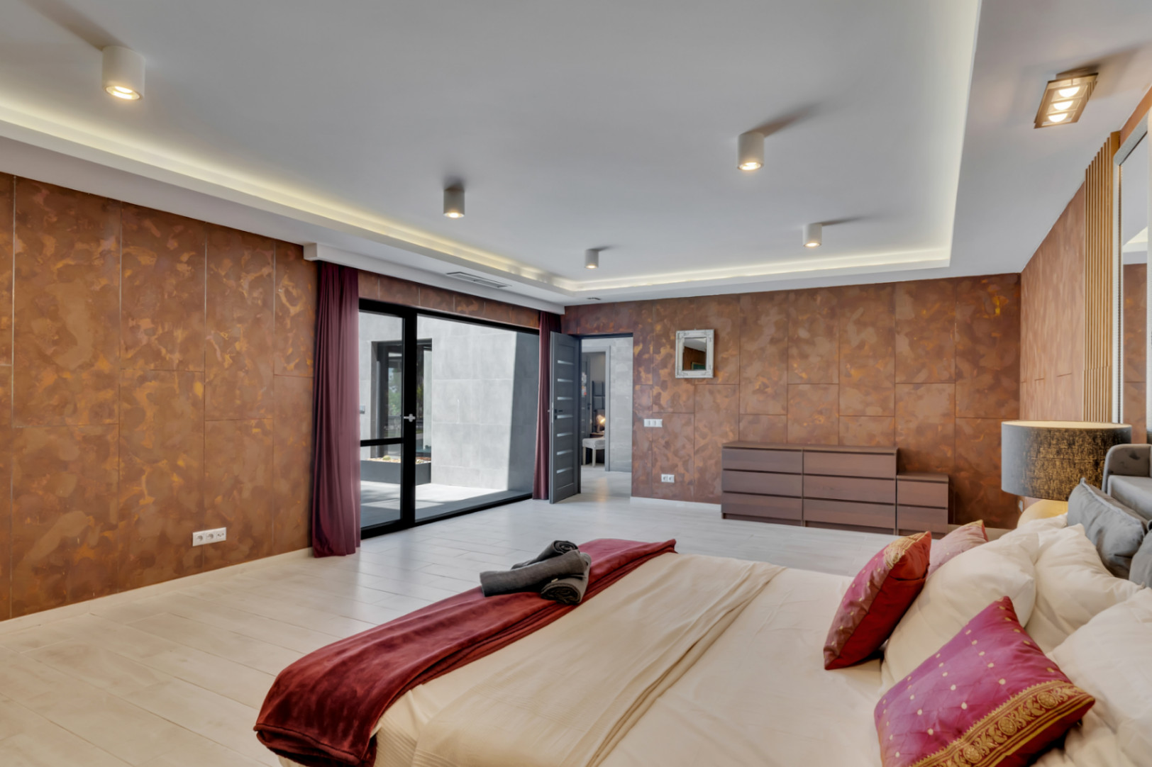 Chambre spacieuse avec murs texturés couleur cuivre et sol clair. Elle bénéficie d'un accès direct à un patio extérieur par une grande porte vitrée coulissante et d'un éclairage de plafond moderne.