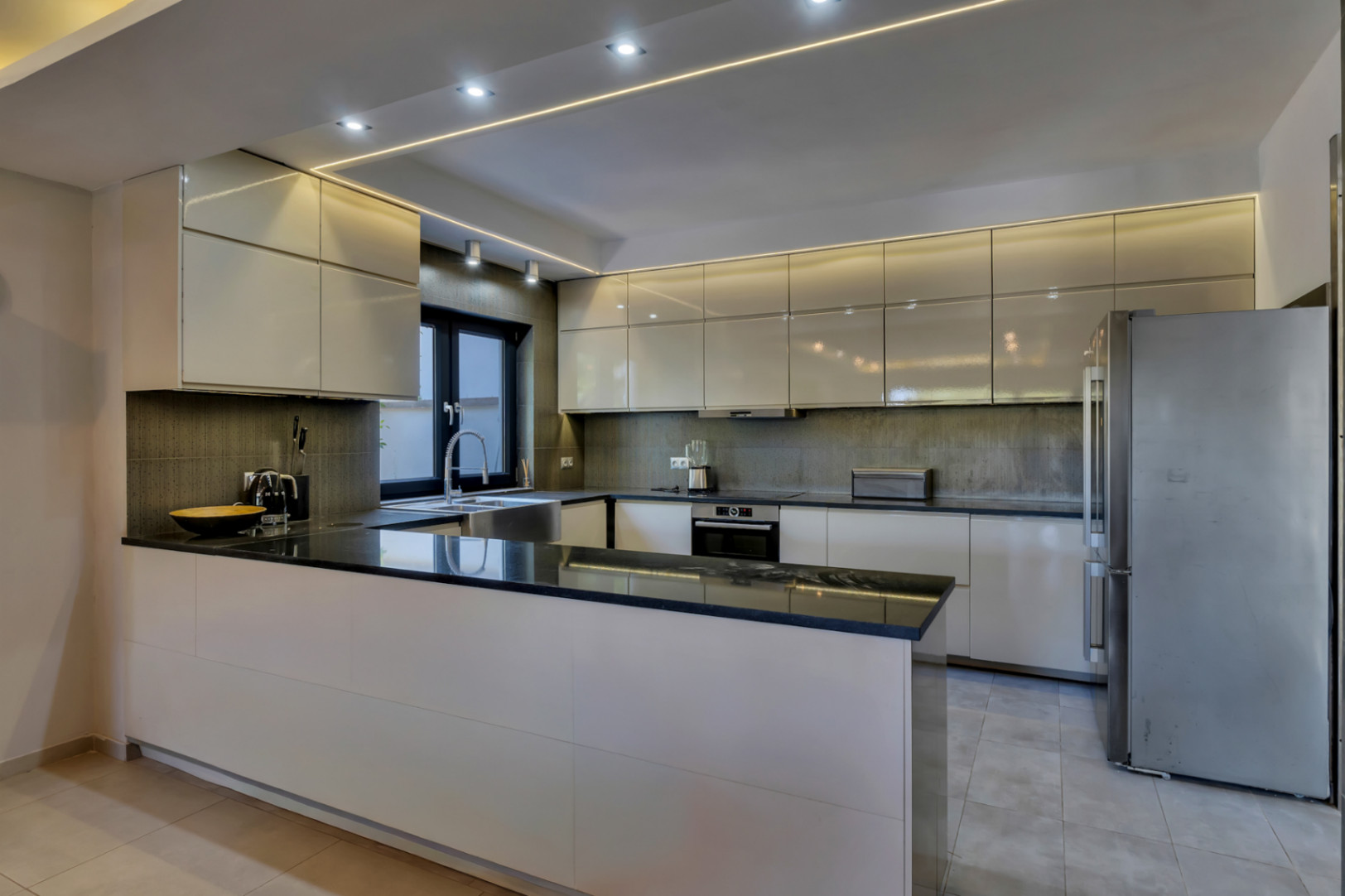 Cuisine moderne avec armoires crème laquées, plans de travail en granit noir et appareils intégrés. Éclairage LED linéaire et encastré au plafond.