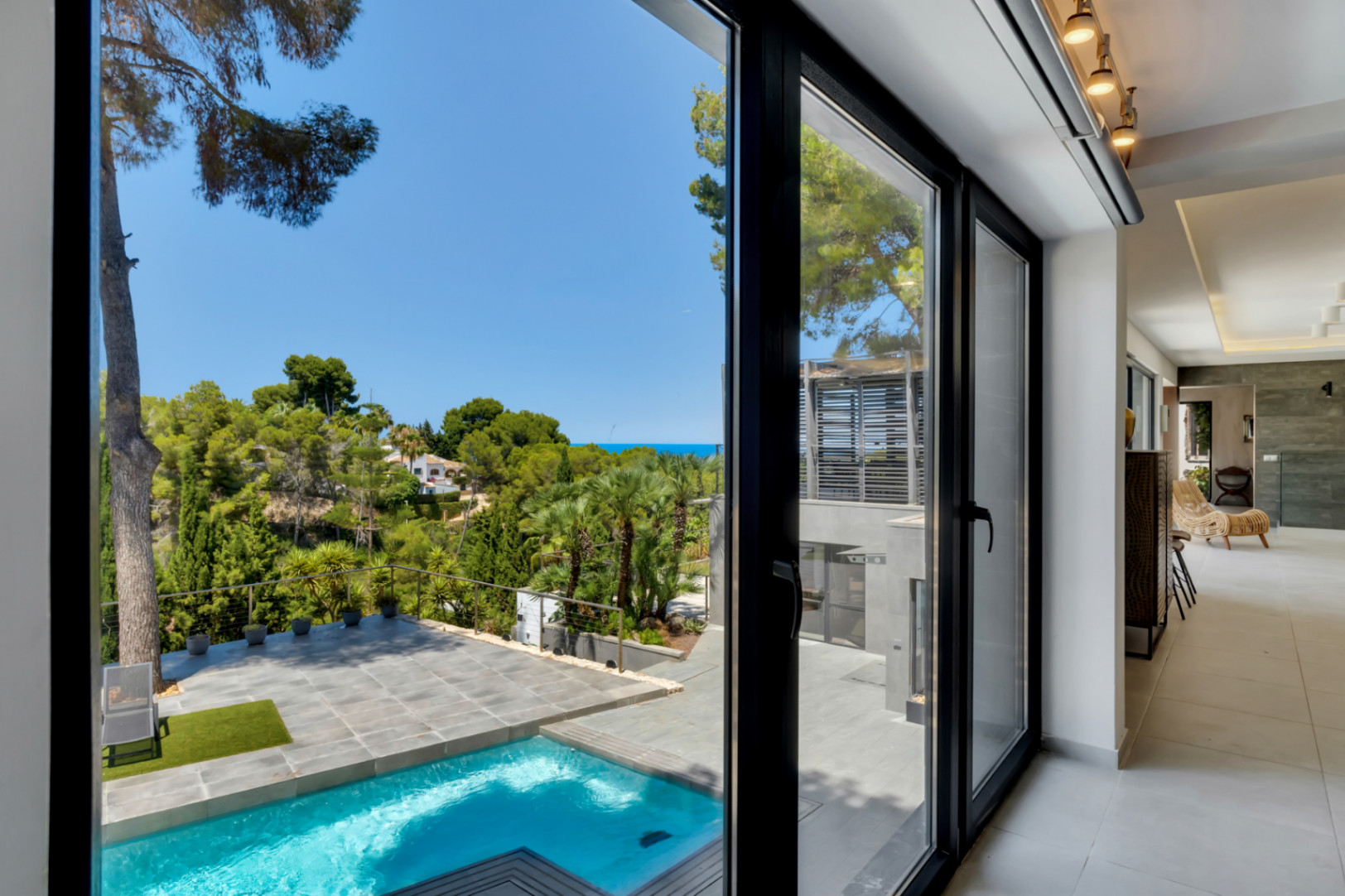 De larges baies vitrées noires relient l'intérieur moderne à la piscine privée, la terrasse carrelée et le paysage méditerranéen luxuriant avec vue sur la mer lointaine.