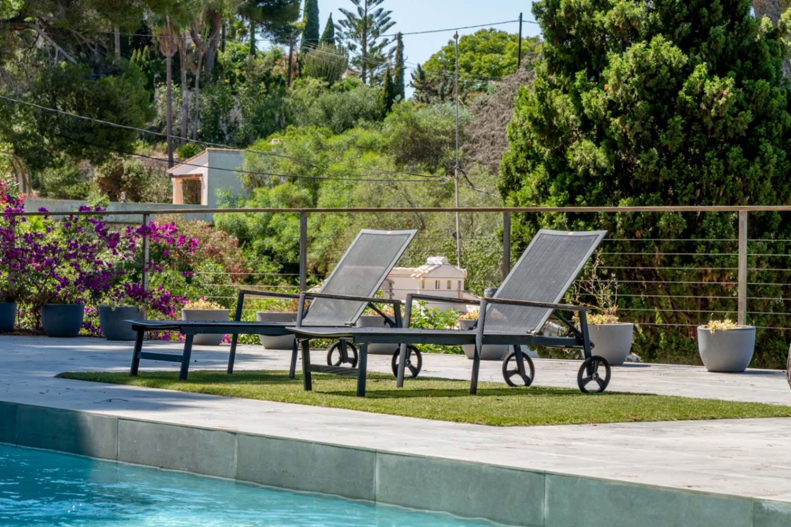 Deux transats contemporains sur gazon synthétique au bord de la piscine, avec une balustrade métallique et une végétation méditerranéenne luxuriante en arrière-plan.