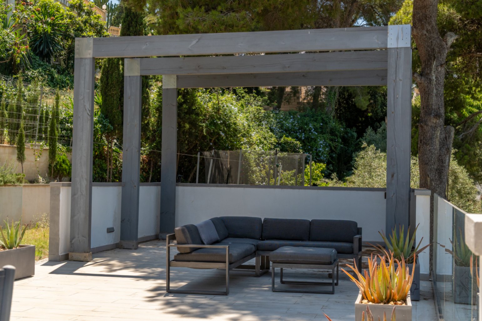 Espace salon extérieur moderne sous une pergola en bois gris, avec canapé d'angle et pouf sur un sol pavé clair, entouré de verdure méditerranéenne luxuriante et de plantes grasses.