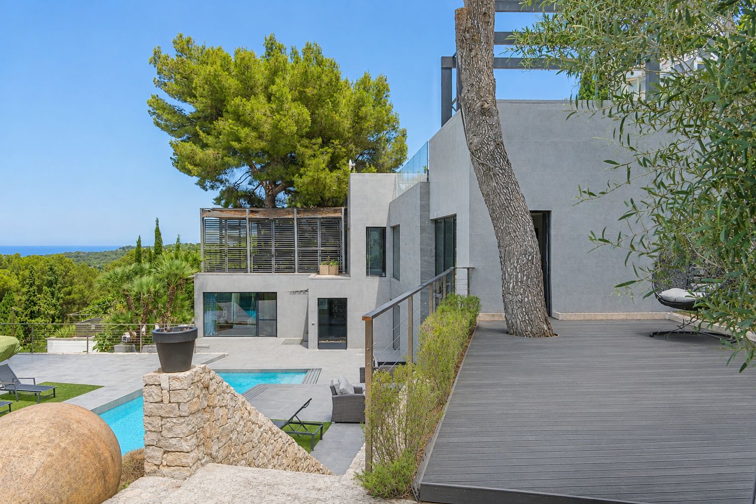 Extérieur de villa moderne à plusieurs niveaux, avec façades grises, piscine privée et terrasse en composite intégrant un arbre. Vues mer lointaines à travers la végétation méditerranéenne.