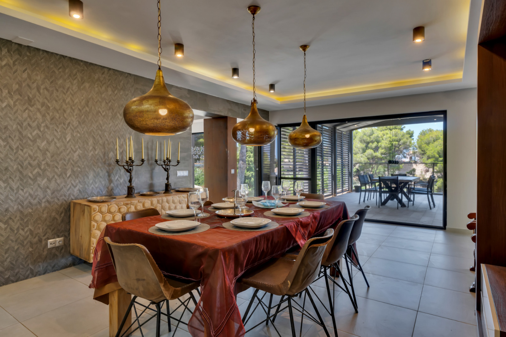 Grande salle à manger avec table en bois et chaises contemporaines, éclairée par trois suspensions dorées et un éclairage indirect. Le mur texturé et le buffet sculpté s'ouvrent sur une terrasse couverte.